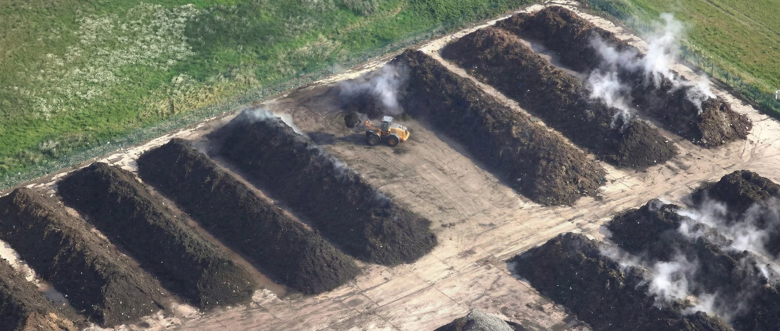 Aerial view of an industrial composting facility with large organic waste windrows and heavy machinery operating between steaming piles.