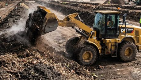 Wheel loader turning steaming compost windrow at an industrial organic waste processing site.