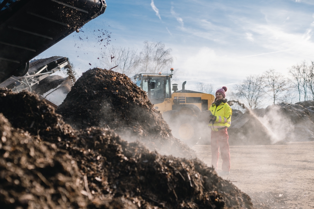 Industrial composting facility with heavy machinery operating near steaming organic waste piles, highlighting dust and airborne exposure risks.