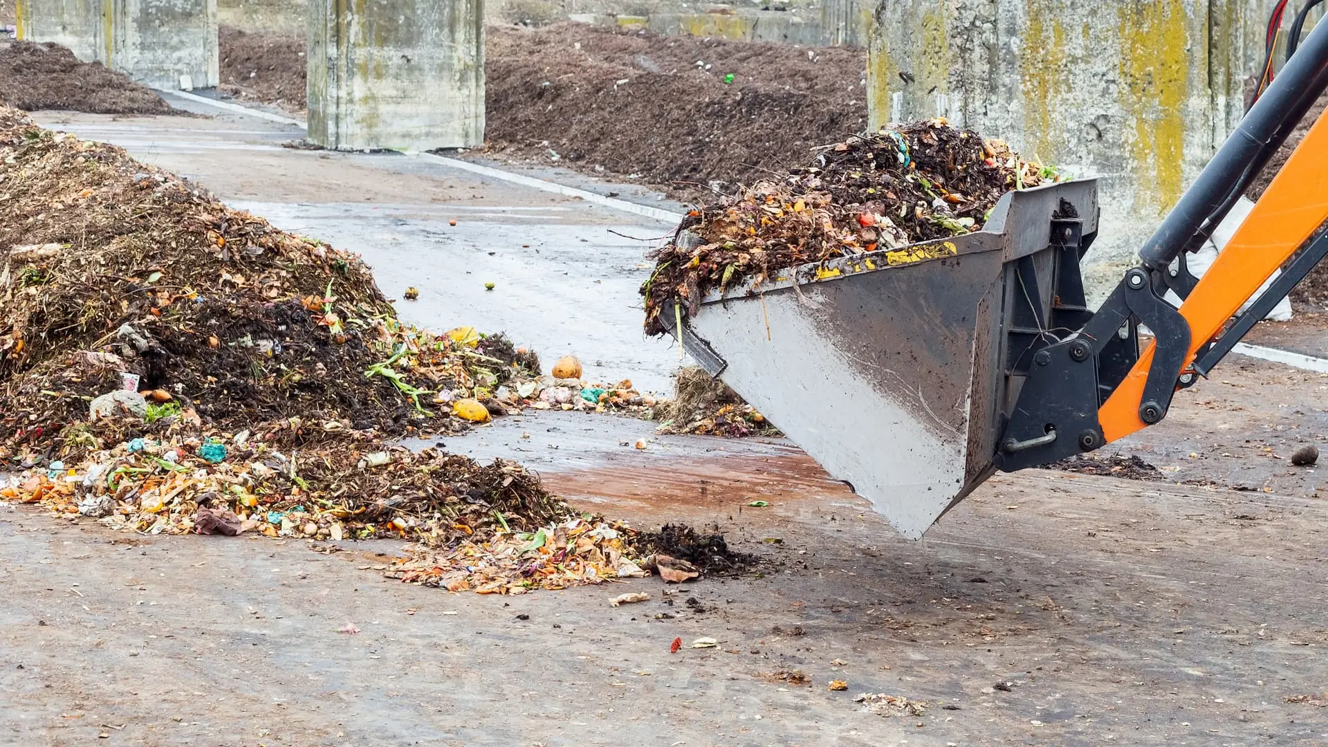 Front-end loader moving decomposing organic waste at a composting facility, with visible food scraps and compost piles on a concrete surface